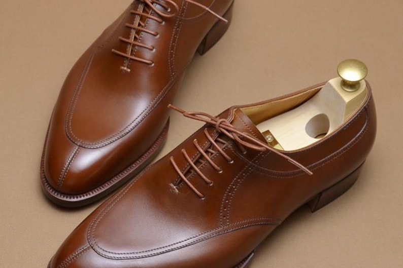 May include: A pair of polished brown leather oxford shoes with a split-toe design and matching brown laces. One shoe features a light-colored wooden shoe tree with a brass-colored knob inserted inside to maintain its shape and structure.