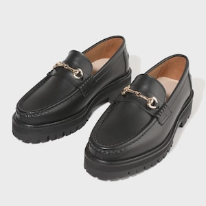 Peut inclure: Une paire de mocassins en cuir noir avec un d&eacute;tail de mors en m&eacute;tal dor&eacute; sur le dessus. Ces chaussures pr&eacute;sentent une semelle &eacute;paisse en caoutchouc crant&eacute; avec un motif &agrave; crampons profonds et un bout arrondi, offrant une allure robuste.
