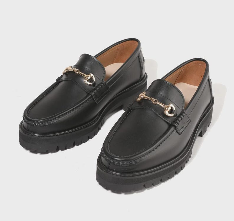 Peut inclure: Une paire de mocassins en cuir noir avec un d&eacute;tail de mors en m&eacute;tal dor&eacute; sur le dessus. Ces chaussures pr&eacute;sentent une semelle &eacute;paisse en caoutchouc crant&eacute; avec un motif &agrave; crampons profonds et un bout arrondi, offrant une allure robuste.