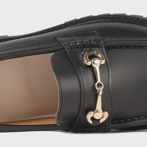 Peut inclure: Un mocassin en cuir noir dot&eacute; d'un d&eacute;tail mors en m&eacute;tal dor&eacute; sur le dessus. La chaussure pr&eacute;sente une semelle &eacute;paisse en caoutchouc crant&eacute; et une doublure int&eacute;rieure beige avec le nom de marque Firenze Atelier imprim&eacute; en blanc sur le talon.