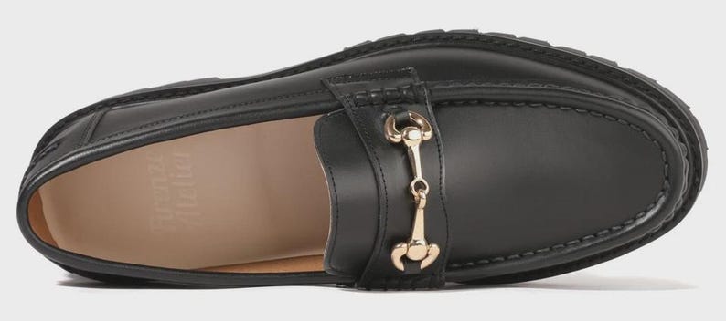 Peut inclure: Un mocassin en cuir noir dot&eacute; d'un d&eacute;tail mors en m&eacute;tal dor&eacute; sur le dessus. La chaussure pr&eacute;sente une semelle &eacute;paisse en caoutchouc crant&eacute; et une doublure int&eacute;rieure beige avec le nom de marque Firenze Atelier imprim&eacute; en blanc sur le talon.