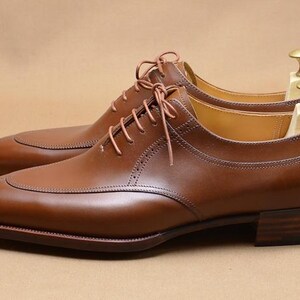 May include: A pair of polished tan leather oxford shoes featuring a split-toe design, thin matching laces, and a stacked wooden heel. Each shoe is equipped with a light-colored wooden shoe tree with a gold-toned metal knob handle at the heel.