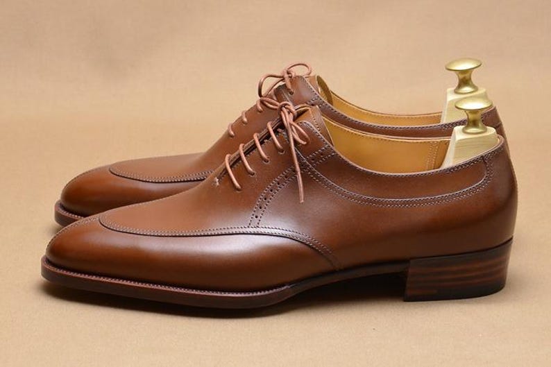 May include: A pair of polished tan leather oxford shoes featuring a split-toe design, thin matching laces, and a stacked wooden heel. Each shoe is equipped with a light-colored wooden shoe tree with a gold-toned metal knob handle at the heel.