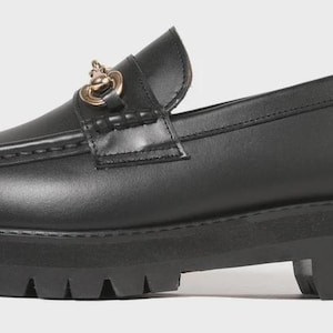 Peut inclure: Un mocassin en cuir noir dot&eacute; d'une &eacute;paisse semelle en caoutchouc crant&eacute;e et d'une cha&icirc;ne en m&eacute;tal dor&eacute; sur la bride sup&eacute;rieure. La chaussure pr&eacute;sente un bout arrondi, des coutures apparentes sur l'empeigne et un contrefort structur&eacute;.