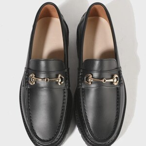 Peut inclure: Une paire de mocassins en cuir noir avec un d&eacute;tail de mors en m&eacute;tal dor&eacute; sur le dessus. Les chaussures pr&eacute;sentent un bout arrondi, une couture apparente le long du bord sup&eacute;rieur et une doublure int&eacute;rieure lisse avec une semelle int&eacute;rieure beige.