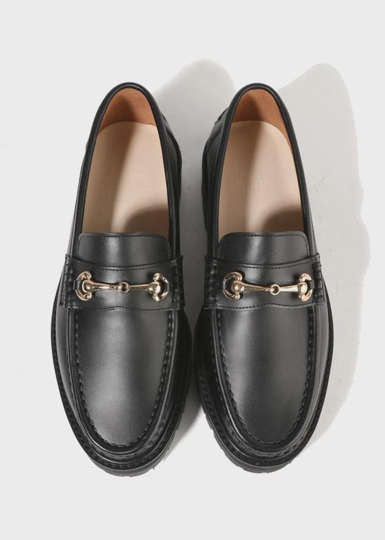 Peut inclure: Une paire de mocassins en cuir noir avec un d&eacute;tail de mors en m&eacute;tal dor&eacute; sur le dessus. Les chaussures pr&eacute;sentent un bout arrondi, une couture apparente le long du bord sup&eacute;rieur et une doublure int&eacute;rieure lisse avec une semelle int&eacute;rieure beige.