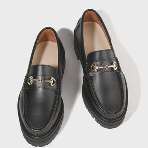 Peut inclure: Une paire de mocassins en cuir noir avec un d&eacute;tail de mors en m&eacute;tal dor&eacute; sur le dessus. Ces chaussures pr&eacute;sentent un bout arrondi, une couture en relief sur l'avant et une &eacute;paisse semelle en caoutchouc noir crant&eacute;e avec un motif antid&eacute;rapant.