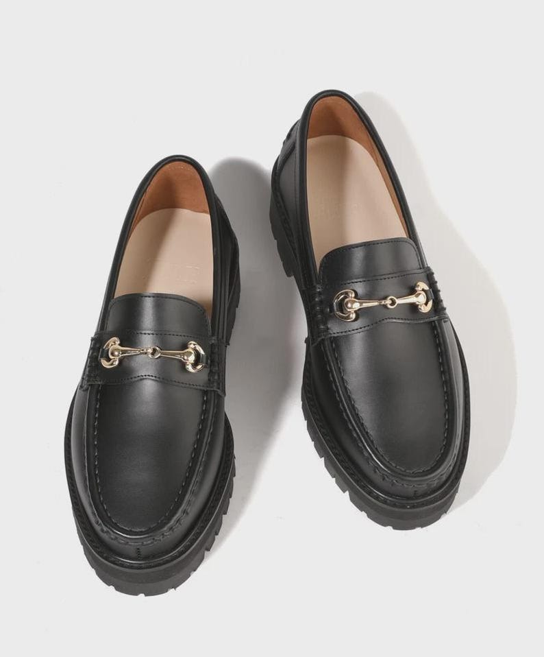Peut inclure: Une paire de mocassins en cuir noir avec un d&eacute;tail de mors en m&eacute;tal dor&eacute; sur le dessus. Ces chaussures pr&eacute;sentent un bout arrondi, une couture en relief sur l'avant et une &eacute;paisse semelle en caoutchouc noir crant&eacute;e avec un motif antid&eacute;rapant.