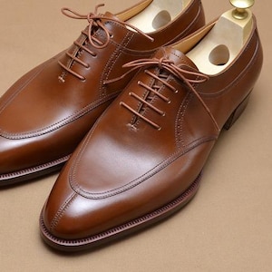 May include: A pair of polished brown leather oxford shoes featuring a split-toe design, tonal stitching, and thin brown laces. Each shoe includes a light-colored wooden shoe tree inserted inside to maintain the shape of the footwear.