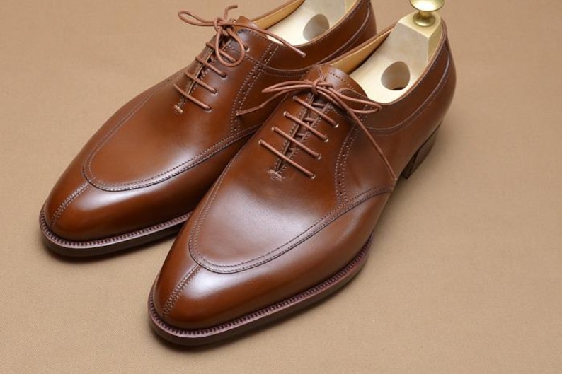 May include: A pair of polished brown leather oxford shoes featuring a split-toe design, tonal stitching, and thin brown laces. Each shoe includes a light-colored wooden shoe tree inserted inside to maintain the shape of the footwear.
