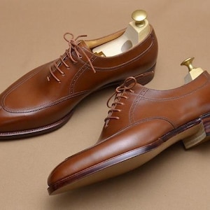 May include: A pair of brown leather oxford shoes with a split-toe design, thin brown laces, and a stacked wooden heel. Each shoe features a light-colored wooden shoe tree inserted inside, with a gold-toned metal knob visible at the top of the heel.