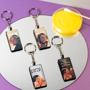 May include: Rectangular keychains with images and text. One keychain says "COME BACK" with a photo of a couple. Another says "I LIKE YOU, YOU PEOPLE... WHATEVER." The other keychains have images of people. The keychains are on a reflective surface.