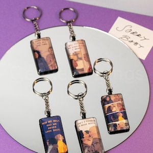 Peut inclure: Porte-clés rectangulaires avec des anneaux argentés. Chaque porte-clés présente une image et un texte différents, dont "Now we walk and we never look back" et "She's a rainbow in beige boots". Les porte-clés sont présentés sur une surface réfléchissante.