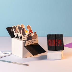 Peut inclure: Une boîte blanche pop-up avec des découpes de personnages et du texte, ainsi que deux blocs noirs avec le texte rouge "KILLING EVE". Une paire de ciseaux et une règle sont au premier plan. Le fond est bleu clair.