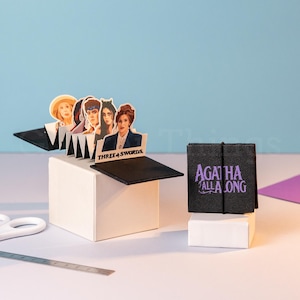 Peut inclure: Une carte pop-up avec des illustrations de personnages et le texte "THREE 4 SWORDS". Un petit livre noir avec le texte "AGATHA ALL ALONG" est également présent. Des ciseaux et une règle sont sur une surface blanche devant un fond bleu.