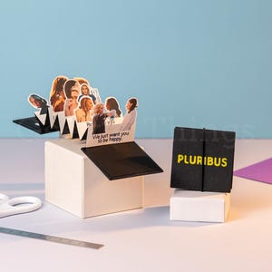Peut inclure: Une carte pop-up avec des images de personnes, le texte "We just want you to be happy !" et une carte noire avec le mot "PLURIBUS" en jaune. Ciseaux et une règle au premier plan.