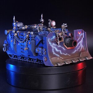May include: A blue armored tank model featuring a front dozer blade painted with white lightning bolts. The vehicle is decorated with metallic chains and small skull ornaments, with a large central cannon and various mounted sensors on top.