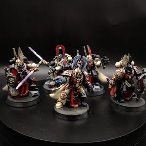Puede incluir: Un grupo de cinco figuras de guerreros en miniatura con armadura negra y crema con detalles en rojo. Cada figura lleva un casco alado, porta un escudo y empuña una espada o un arma de fuego. Los soldados están sobre bases negras circulares.