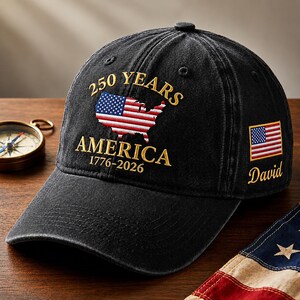 May include: A black baseball cap featuring embroidered gold text that reads 250 YEARS AMERICA 1776-2026 above a red, white, and blue map of the United States. The side of the cap displays a small American flag patch and the embroidered name David.