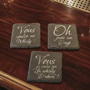 Peut inclure: Trois dessous de verre carrés en ardoise avec une écriture blanche. Les dessous de verre portent des phrases françaises : "Vous voulez un Whisky ?", "Oh juste un Doigt" et "Vous ne voulez pas Un whisky D'abord ?" Les dessous de verre sont sur une surface en bois foncé.