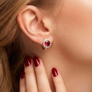 May include: A gold earring featuring a central red gemstone encircled by smaller white gemstones. The earring is worn on an ear. The person's nails are painted a deep red.