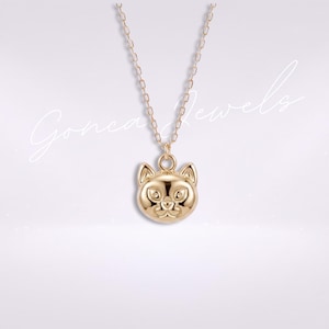 Puede incluir: Un collar de tono dorado con un colgante en forma de cabeza de gato con orejas puntiagudas y rasgos faciales definidos. El colgante cuelga de una cadena delicada. El texto Gonca Jewels está escrito en letra cursiva sobre la imagen.