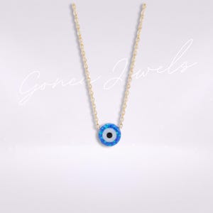 May include: A gold chain necklace featuring a circular evil eye pendant with a blue opal-like finish, a white center, and a black pupil. The text Gonca Jewels is overlaid in white script across the center of the image.
