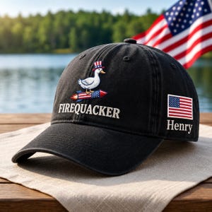 May include: Black baseball cap with the embroidered text "FIREQUACKER" and a cartoon duck riding a rocket. Features an American flag patch and the name "Henry". The cap is on a wooden surface with an American flag in the background.