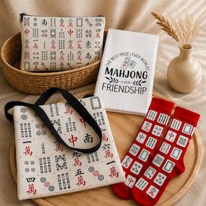 Könnte beinhalten: Eine Sammlung von Accessoires zum Thema Mahjong, darunter eine Tragetasche, eine Kosmetiktasche, Socken und ein Küchentuch. Die Artikel zeigen Mahjong-Kachel-Designs in Rot, Schwarz und Grün auf cremefarbenem Hintergrund. Das Tuch trägt den Text "The best prize I ever won at Mahjong is our friendship."