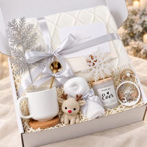 Op de afbeelding: Een geschenkdoos met een witte gebreide deken, een geribbelde witte mok op een kurken onderzetter, een gouden lepel, een klein gehaakt rendier, een witte kaars met het label Smells like Ever, een witte handdoek en een kaart met Best Wishes.