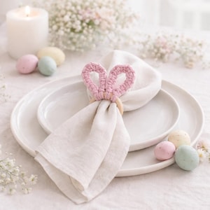 Peut inclure: Une serviette en lin blanche est maintenue par un anneau en bois avec des oreilles de lapin en crochet rose. La serviette est posée sur une assiette blanche, avec des œufs de Pâques pastel à proximité. Une bougie allumée et des fleurs sont en arrière-plan.