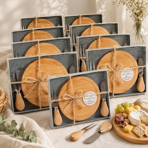 May include: A collection of boxed cheese board sets, each containing a round bamboo board and two metal cheese knives with wooden handles. The sets are tied with twine and feature a circular sticker that reads Thank you for celebrating with us.