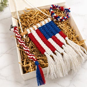 May include: A patriotic gift set featuring a macrame American flag wall hanging with red, white, and blue stripes and tassels. Includes a matching bracelet and a twisted rope ornament with a tassel, all in a gift box.