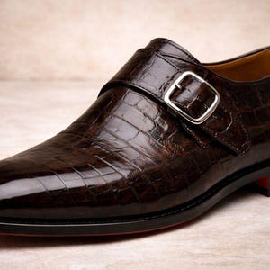 May include: A single, brown leather shoe with a crocodile skin texture. The shoe features a silver buckle closure and a black sole with a red accent. The shoe is a formal style.