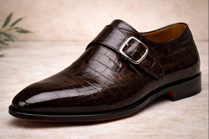 May include: A single, brown leather shoe with a crocodile skin texture. The shoe features a silver buckle closure and a black sole with a red accent. The shoe is a formal style.