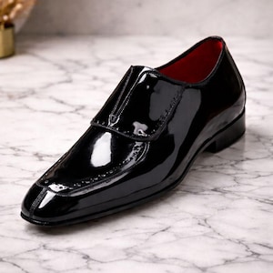 May include: A glossy black leather shoe with a red interior. The shoe features a sleek design with a pointed toe and decorative stitching along the edges. The shoe is a formal style.