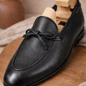 May include: A black leather loafer with a bow detail. The shoe has a textured surface and a wooden shoe tree inside. The loafer is displayed on a wooden surface.