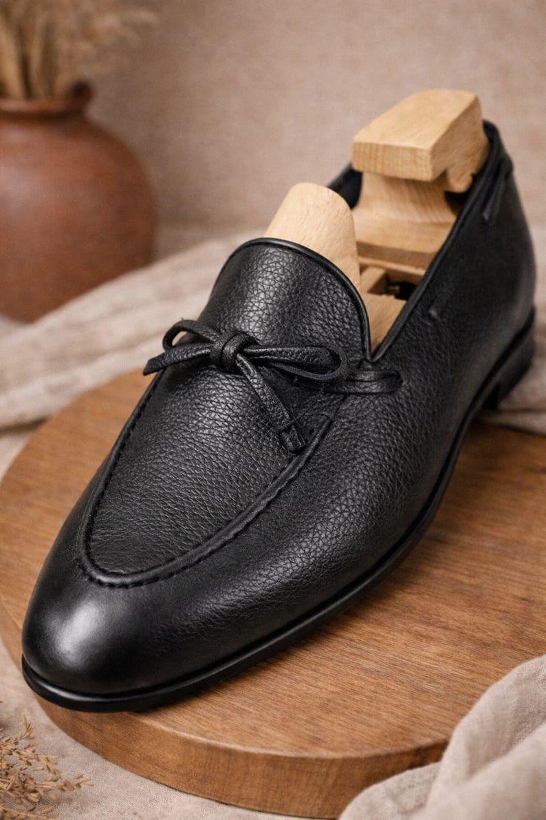 May include: A black leather loafer with a bow detail. The shoe has a textured surface and a wooden shoe tree inside. The loafer is displayed on a wooden surface.