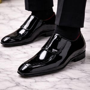May include: Close-up of a pair of shiny black patent leather dress shoes. The shoes have a sleek, polished appearance with a glossy finish. The shoes are worn with black socks and trousers. The shoes have a decorative design on the toe.