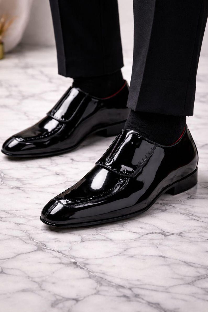 May include: Close-up of a pair of shiny black patent leather dress shoes. The shoes have a sleek, polished appearance with a glossy finish. The shoes are worn with black socks and trousers. The shoes have a decorative design on the toe.