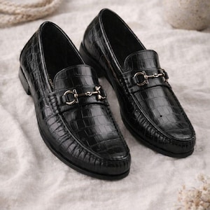May include: A pair of black leather loafers with a crocodile texture. Each shoe features a silver-colored metal ornament on the vamp. The shoes are displayed on a white, textured surface.