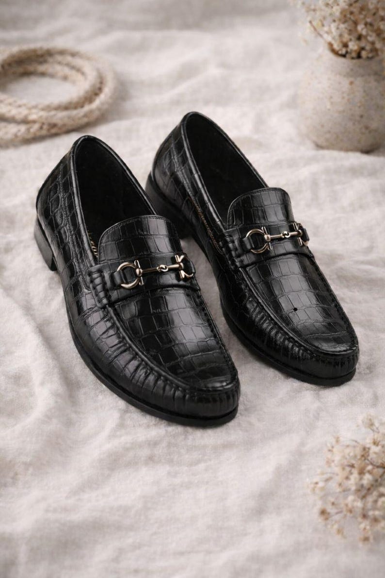 May include: A pair of black leather loafers with a crocodile texture. Each shoe features a silver-colored metal ornament on the vamp. The shoes are displayed on a white, textured surface.