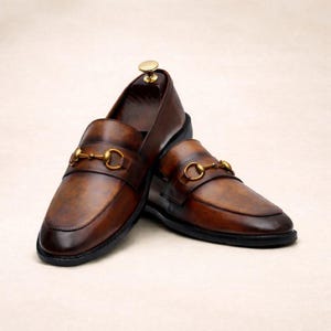 Puede incluir: Un par de mocasines de cuero marr&oacute;n con un detalle de adorno met&aacute;lico dorado. Los zapatos tienen un dise&ntilde;o cl&aacute;sico con punta redondeada y tac&oacute;n bajo. El cuero tiene un acabado envejecido.