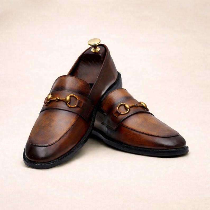 Puede incluir: Un par de mocasines de cuero marr&oacute;n con un detalle de adorno met&aacute;lico dorado. Los zapatos tienen un dise&ntilde;o cl&aacute;sico con punta redondeada y tac&oacute;n bajo. El cuero tiene un acabado envejecido.