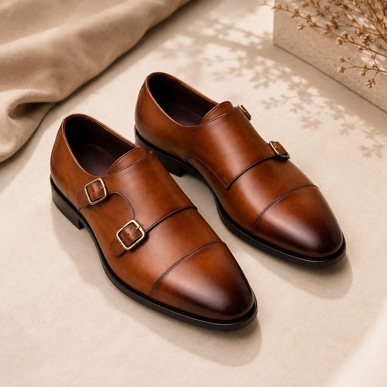 May include: A pair of brown leather double monk strap shoes featuring a cap toe design, two gold-toned metal buckles on each side, and a dark brown sole. The leather has a polished finish with a subtle gradient from light to dark brown.
