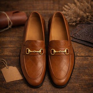 May include: A pair of brown leather loafers with a diamond pattern and gold-colored metal accents. The shoes are displayed on a wooden surface, showcasing their classic design and craftsmanship. The loafers have a slip-on style.