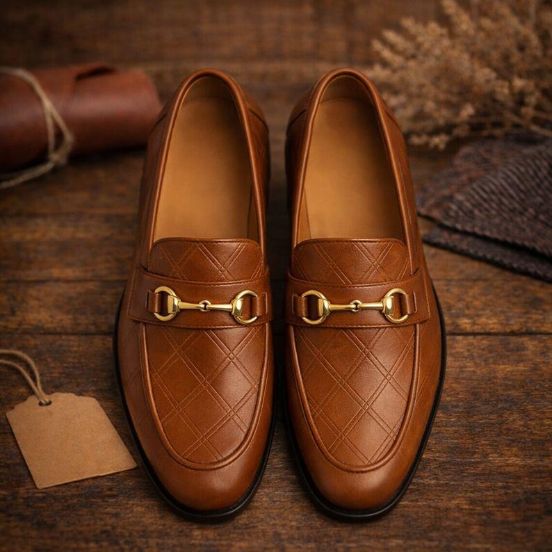 May include: A pair of brown leather loafers with a diamond pattern and gold-colored metal accents. The shoes are displayed on a wooden surface, showcasing their classic design and craftsmanship. The loafers have a slip-on style.