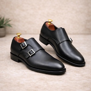 May include: A pair of black leather double monk strap shoes. The shoes have silver buckles and are displayed on a neutral background. The shoes are on wooden shoe trees.