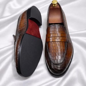 K&ouml;nnte beinhalten: Ein Paar braune Leder-Loafer mit Krokodilleder-Textur. Die Schuhe haben eine dunkle Sohle und ein rotes Wildleder-Innenfutter. Die Schuhe werden auf einer wei&szlig;en Satin-Oberfl&auml;che pr&auml;sentiert.