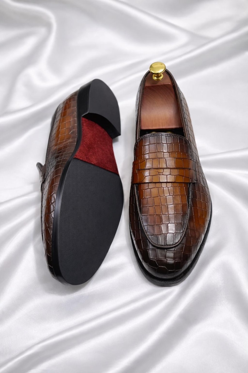 K&ouml;nnte beinhalten: Ein Paar braune Leder-Loafer mit Krokodilleder-Textur. Die Schuhe haben eine dunkle Sohle und ein rotes Wildleder-Innenfutter. Die Schuhe werden auf einer wei&szlig;en Satin-Oberfl&auml;che pr&auml;sentiert.